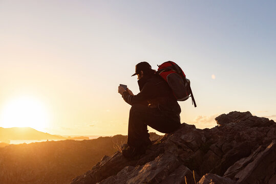 Side view of unrecognizable male hiker sitting on rock in highlands and taking photo while admiring sunset