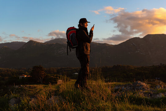 Side view of hiker standing on rock in highlands and speaking on cellphone while admiring sunset