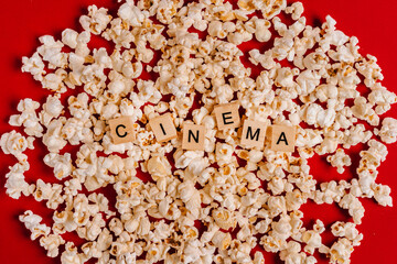 The word "CINEMA" made of wooden blocks lies on popcorn on a red background. The concept of recreation with watching movies