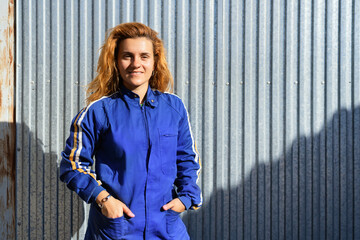 Cheerful female engineer in protective glasses and workwear standing near metal wall at plant and looking at camera while enjoying break during work