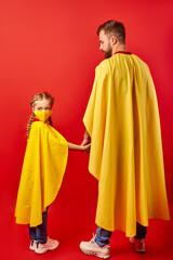 Obraz premium father and daughter ready to fight against coronavirus, they stand in superhero costume, girl in medical mask, look at camera. isolated over red background