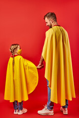 Fototapeta premium friendly man and kid girl in yellow cloaks, pretend to be superheroes. male and child in mask stand holding hands together, rear view