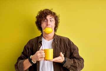 young male stuffed his mouth with lemon, want to be healthy. hold cup in hands isolated over yellow...