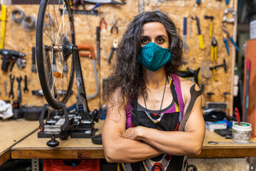 Serious adult ethnic female mechanic in workwear and protective mask holding wrench and looking at camera while standing against weathered workbench with various tools in bicycle service workshop