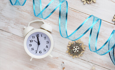 White alarm clock showing 12 o'clock in the day or night and decorative blue ribbon on a wooden background. Christmas and new year's composition.
