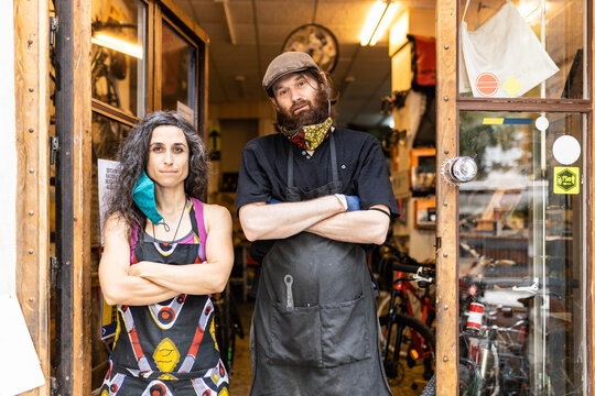 Confident adult ethnic man and woman in workwear with protective masks standing on doorway of bicycle service workshop and looking at camera