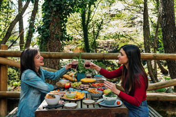 Side view of friendly women sitting at table in woods and enjoying delicious food during picnic in Valle del Jerte while talking and looking at each other