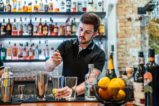 Serious male bartender preparing cocktail with alcohol beverage and ice cubes while standing at counter in bar