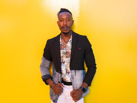 Serious African American male model in fancy outfit standing near yellow wall on street and looking at camera