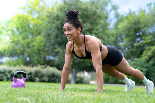 Strong African American female athlete doing push ups on green lawn in park while smiling and looking away