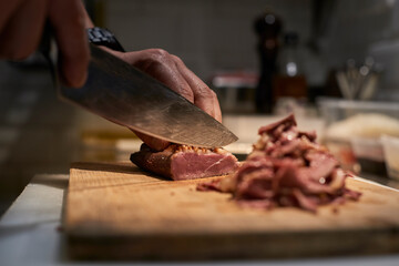 Anonymous chef standing at table in kitchen and cutting palatable smoked meat while preparing dish in restaurant