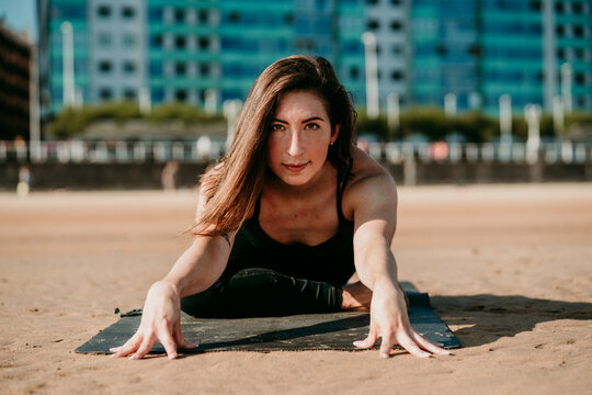 Smiling flexible female sitting on mat in Janu Sirsasana and doing forward bend while looking at camera and practicing yoga on beach in summer