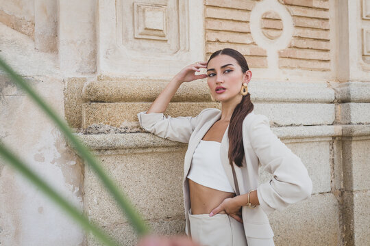 Confident young ethnic female in fashionable beige suit and white top leaning elbow on stone wall of urban building and looking away
