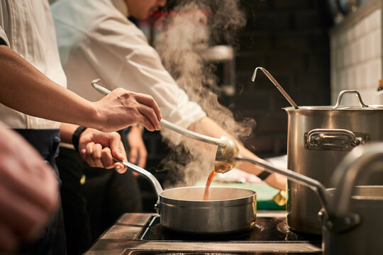 Side view of unrecognizable chef in uniform preparing delicious sauce while standing at stove in kitchen of restaurant