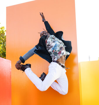 Low angle of carefree African American male in fancy outfit in moment of jumping above ground near vibrant orange wall in city