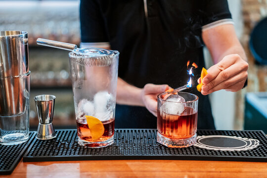 Unrecognizable male bartender preparing Negroni cocktail and setting orange slice on fire while standing at counter