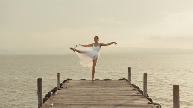 Full body young graceful female dancer in white gown standing on tiptoes with leg raised while performing  ballet movements on wooden boardwalk against sea in summer evening