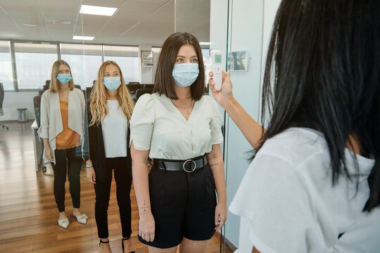 Receptionist with thermometer gun checking body temperature of coworkers before entering office space during coronavirus pandemic