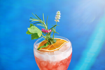 Glass of delicious cold fruit drink with orange slice and herbs placed at poolside against blue water of swimming pool in summer day
