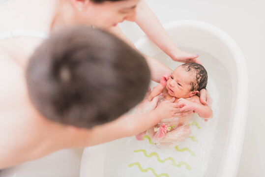 From above unrecognizable parent and kid washing crying newborn baby in warm water in basin at home