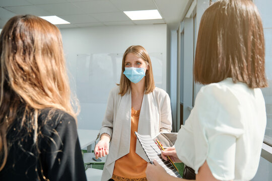 Young female manager in medical mask explaining business idea to coworkers while standing together in modern workspace and discussing project