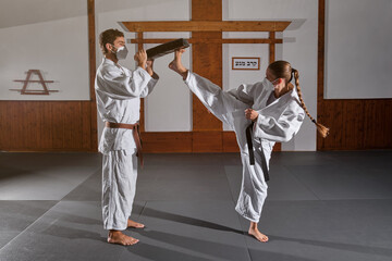 Woman with braid wearing protective mask and white kimono with a black belt attached kicking a man's mitten into the air