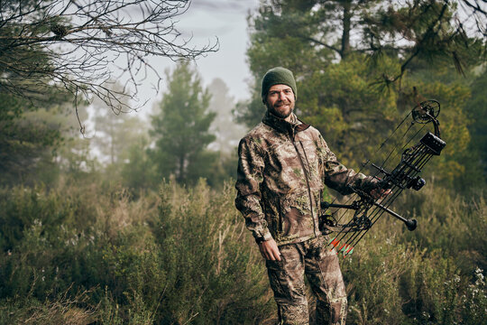 Cheerful man in camouflage standing with compound bow in forest and looking at camera during hunting