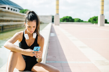 Fit female with tattoo on arm and headphones on neck dressed in black sports bra and shorts browsing mobile phone while sitting on stone border and relaxing after fitness workout on street