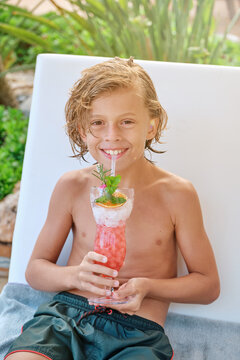 Cheerful preteen boy with wet curly hair sitting on chair and enjoying refreshing fruit drink while spending summer day at poolside in resort