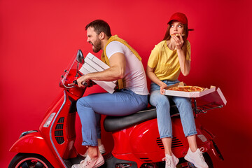 Obraz na płótnie Canvas funny redhead woman rides with courier boyfriend delivering food orders on motorcycle, female is eating pizza sitting behind man, isolated red background