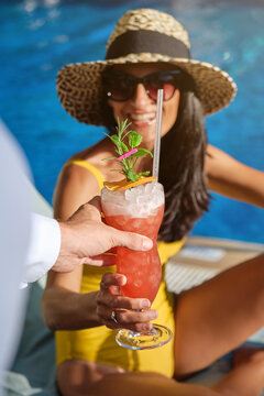 Fototapeta Cheerful young female tourist in stylish straw hat and sunglasses receiving fresh cold fruit beverage from unrecognizable barmen while enjoying summer vacation at poolside in resort