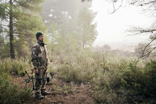 Side view of man in camouflage standing with compound bow in forest and looking away during hunting