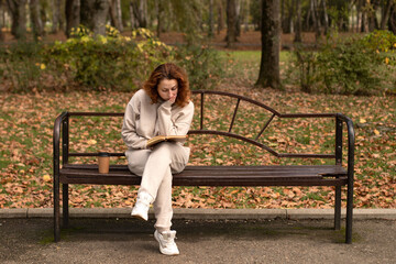 Naklejka na ściany i meble a young beautiful girl is sitting on a bench in an autumn Park and reading a book