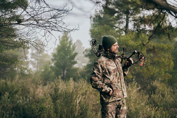 Side view of man in camouflage standing with compound bow in forest and looking away during hunting