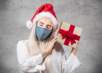 Santa woman wearing face mask and holding christmas gift isolated on gray background. Happy young girl wearing red santa hat and holding present box. A Corona - Covid 19 Christmas concept