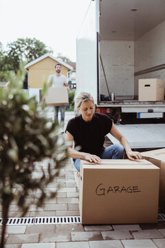 Woman crouching by cardboard box while partner unloading luggage from moving van
