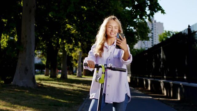Blonde hair woman in the middle of the street make a stop with her electric scooter to make some pictures with the smartphone she smiling large and feeling very happy