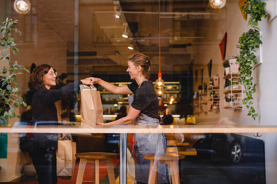 Side view of smiling saleswoman giving shopping bag to female customer at checkout counter seen through glass window