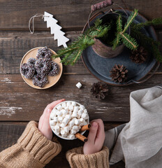 Hands keep christmas mug of cacao and marshmallow with cinnamon on an old brown wooden table with cookies, cones and fir.