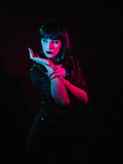 Fototapeta na wymiar Girl with black hair and black makeup posing on a black background with colored light. She demonstrates her arms and stares to the side.