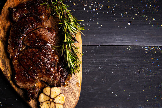 fried beef steak drizzled with butter lies on a wooden cutting board with rosemary, chili and garlic on a black background. flat lay. Horizontal photo. free space for advertising logotypes of inscript