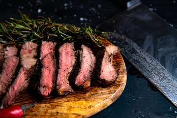 a piece of a beautiful juicy fried steak is cut and lies on a board with rosemary and chili on a black background close-up and a butcher's knife lies next to it. Horizontal photo