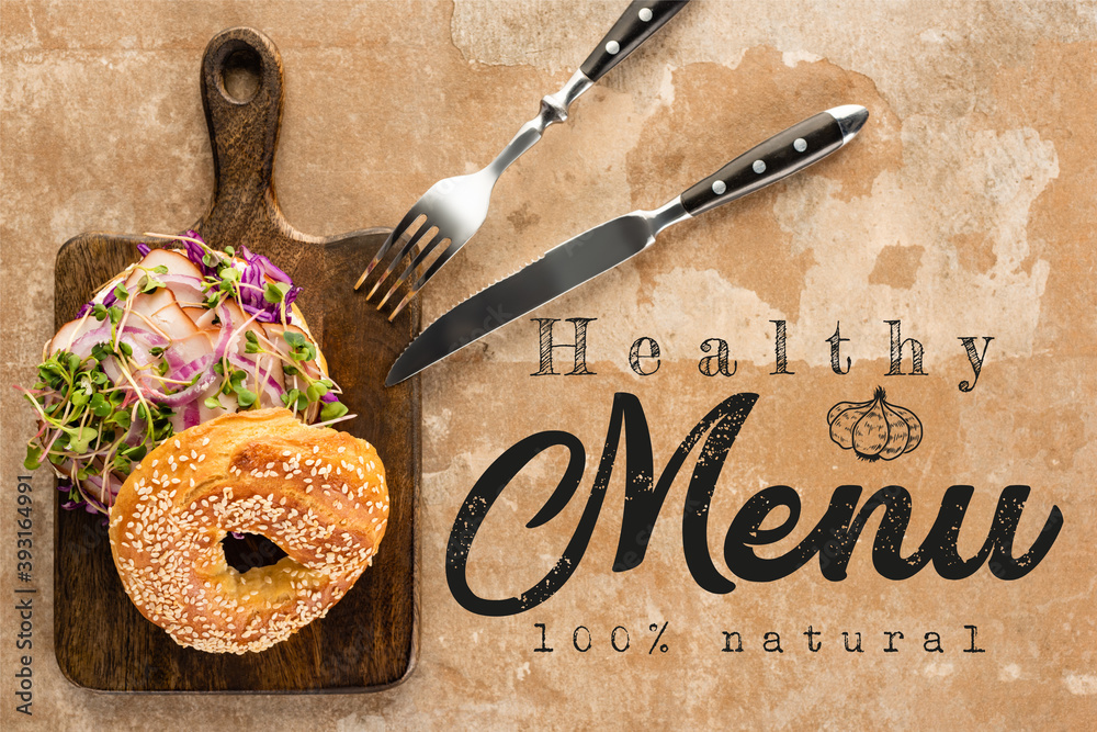 Wall mural top view of bagel with meat on cutting board with cutlery near the healthy menu lettering on texture