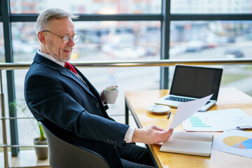 Businessman checks a printed report or document when he sits, working on paper work at a laptop.