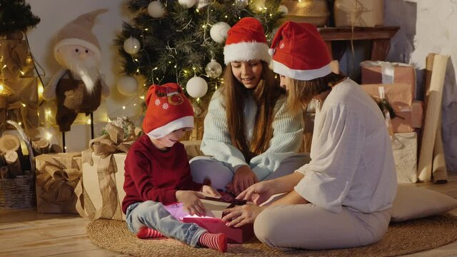 Mom with children in Santa hats sitting on the carpet near the Christmas tree and decorations open a gift with light inside