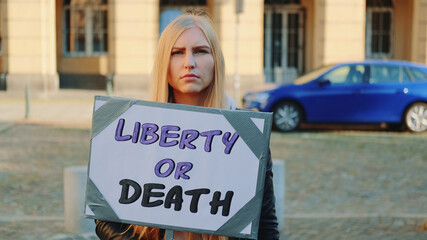 Blonde woman with protest banner calling to choose liberty or death. She walking on the street.
