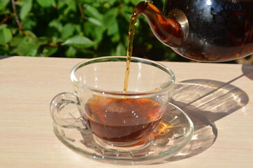 A girl pours tea from a transparent teapot into a transparent cup against a background of green foliage Good morning, outdoor cafe, energy boost. Tea being poured into glass tea