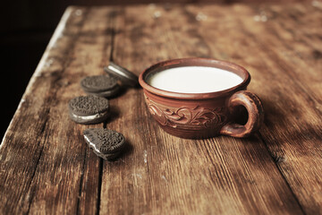There is a bouquet of lilies of the valley on a wooden table, milk and chocolate chip cookies in a clay cup.
