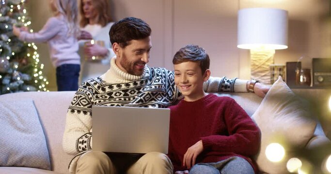 Happy Caucasian dad with teen son sitting in cozy room typing on laptop spending holiday time on internet buying gifts. Mom and small kid decorating glowing Christmas tree. Family holidays concept