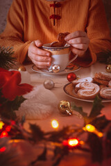 Lady in an orange sweater drinks tea with puff cookies among Christmas decorations and Santa's hats